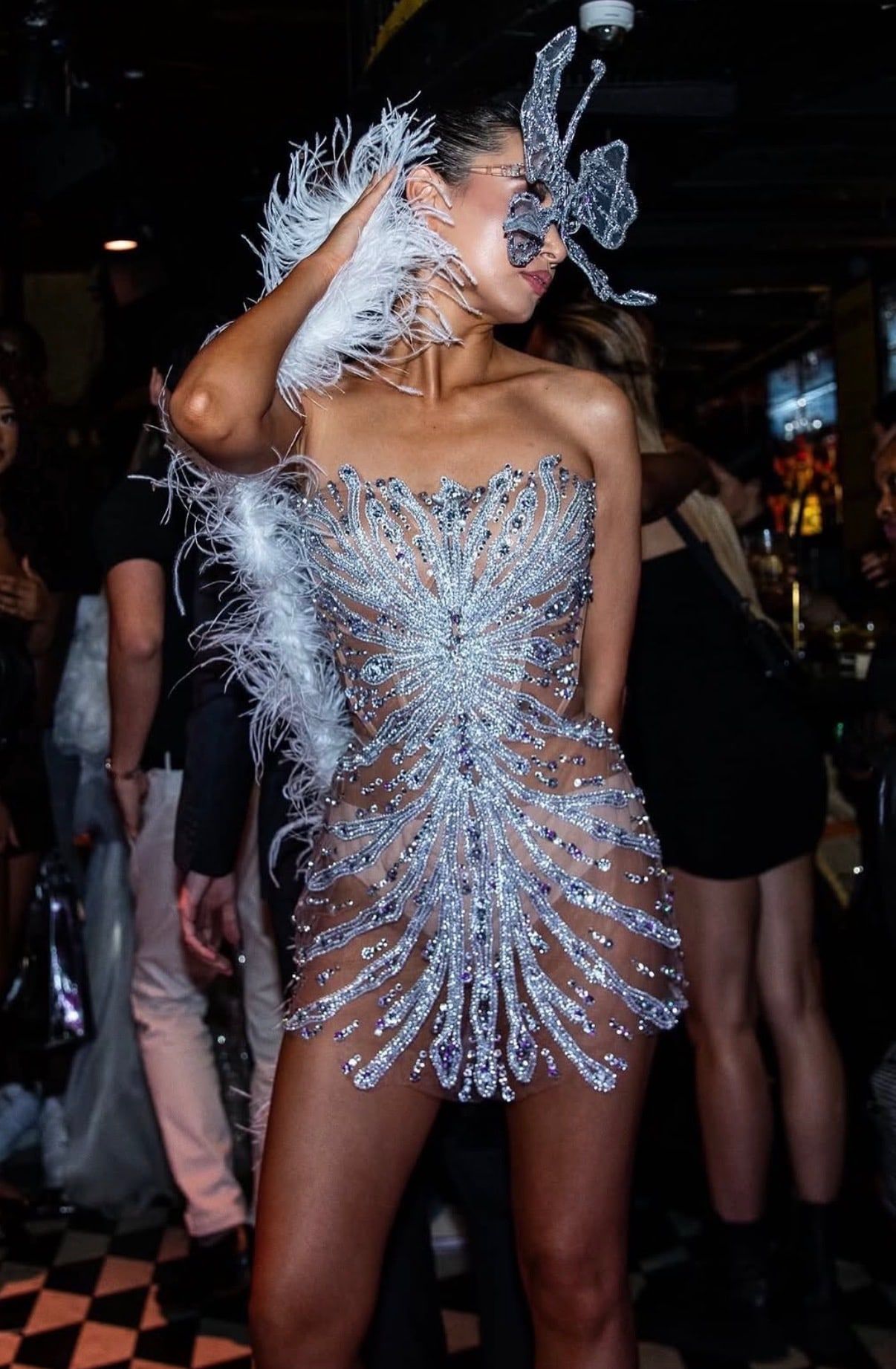 A model in a sheer crystal-embellished mini dress, a white feather boa and a crystal butterfly mask