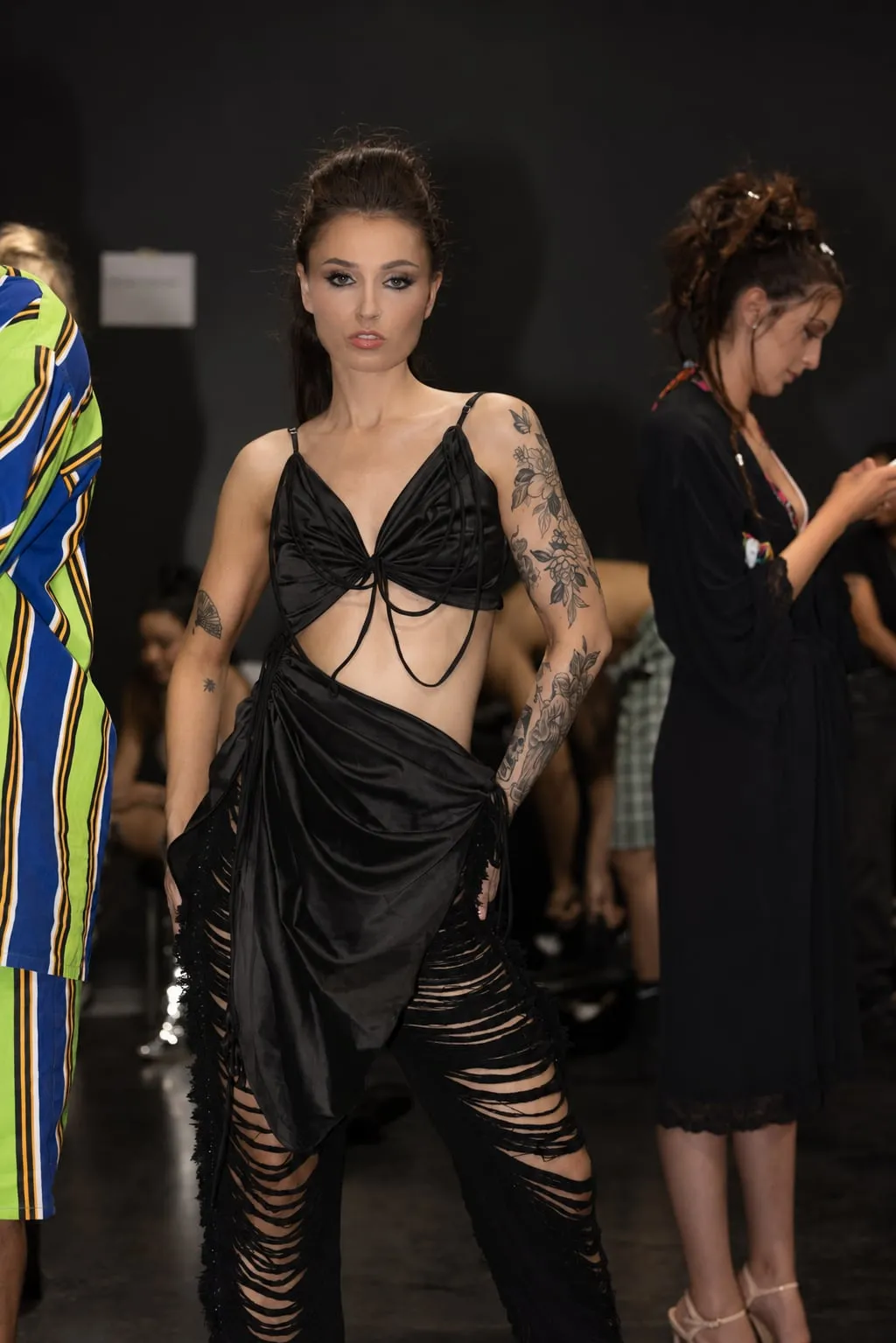 A tattooed model in a black draped top and shredded trousers backstage