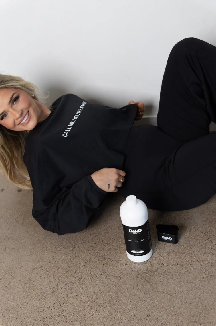A smiling woman lying on the studio floor beside a liter bottle of BakD Sunless solution and an application mitt