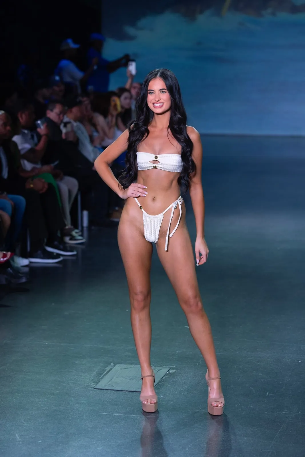 A model with long dark hair in a white bikini on the runway in front of the audience